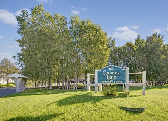 Sign reading "810 Country Lane Apartment Homes" stands on a lush, green lawn with a backdrop of tall trees under a clear blue sky, conveying tranquility.