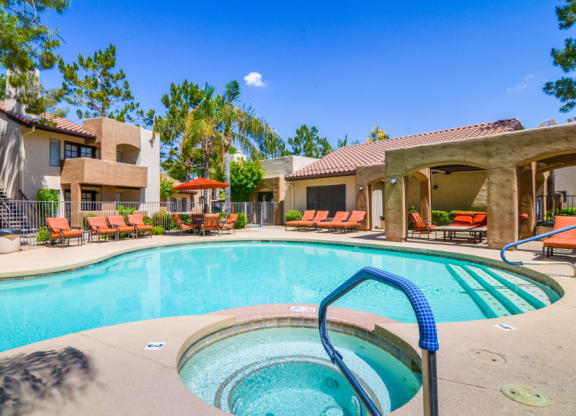 Sunny outdoor pool area here at Desert Club Apartments with clear blue water, surrounded by orange lounge chairs and shaded cabanas. Lush palm trees and a clear sky create a tranquil vibe.