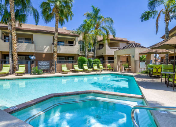 Sunny pool area here at Desert Horizon Apartments with clear blue water, surrounded by green lounge chairs and tall palm trees. Beige apartments in the background and a relaxed, inviting atmosphere.
