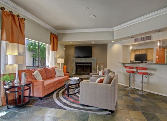 A modern living room here at Desert Horizon Apartments with a gray slate floor, orange and beige sofas, and large windows with orange curtains. A fireplace has a TV above it. Two red bar stools are at a kitchen counter, adding a cozy, contemporary feel.