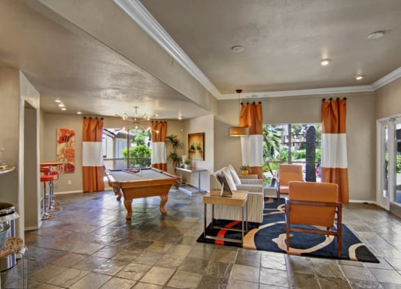 Spacious living area here at Desert Horizon Apartments with a pool table, gray tile floor, and orange-accented decor. Bright with natural light from large windows. Modern and inviting.