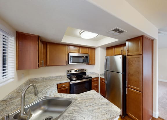 Modern kitchen here at Desert Horizon Apartments with stainless steel appliances, light brown wooden cabinets, granite countertops, and a double sink. Adjacent is a carpeted room.