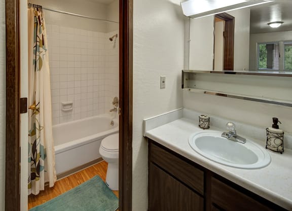 Bathroom here at Driftwood Apartments with a white-tiled bathtub and shower curtain, wooden floor, and vanity sink. Soft lighting and a calm, clean atmosphere.