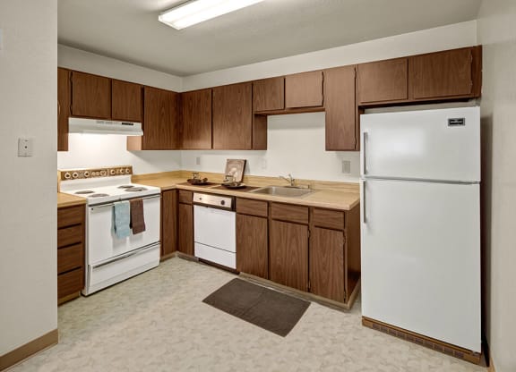 Kitchen here at Driftwood Apartments with wooden cabinets, white appliances, and hardwood floor. Includes stove, refrigerator, dishwasher, and sink. Clean, tidy, and functional space.