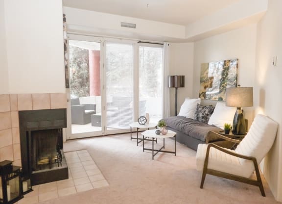 Modern living room here at Cheyenne Creek Apartment Homes with a cozy fireplace, white couch, and armchair. Natural light from large windows illuminates a serene decor and artwork.
