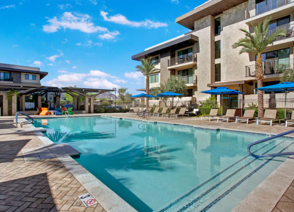 Modern Escape Apartments courtyard with a tranquil swimming pool, surrounded by palm trees and lounge chairs. Blue umbrellas offer shade under a bright sky.