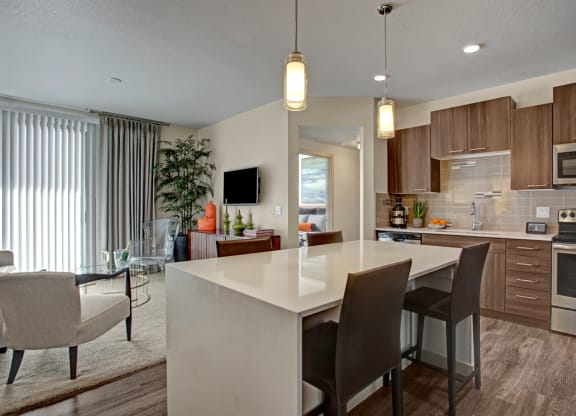 Modern kitchen and living area here at Escape Apartments with brown cabinets, stainless steel appliances, a white island with dark chairs, and cozy seating. Elegant and inviting ambiance.