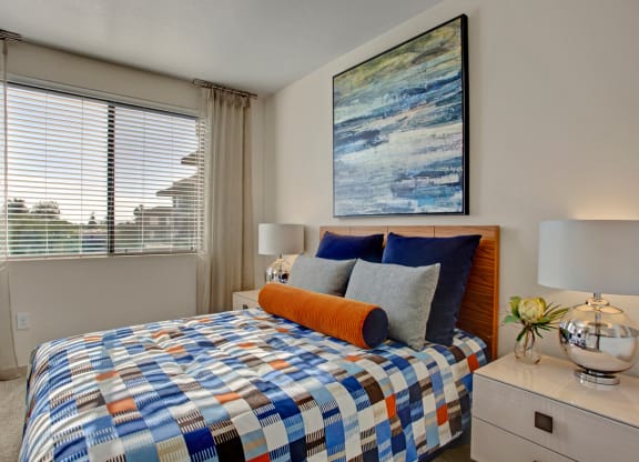 Modern bedroom here at Escape Apartments with a colorful patchwork bedspread, navy and gray pillows, and an orange bolster. Abstract wall art and a silver lamp add elegance.