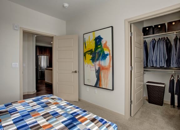 Bedroom here at Escape Apartments with abstract art and open closet. Closet contains neatly arranged blue shirts. Bedspread features a colorful geometric pattern. Neutral tones create a calm atmosphere.