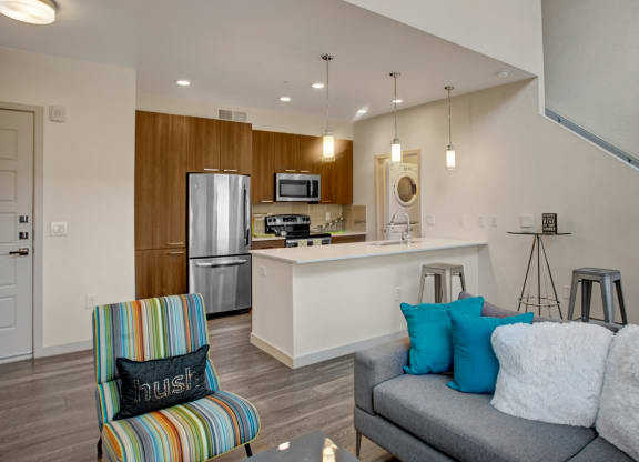Modern living room and kitchen here at Escape Apartments with open layout. Grey sofa with blue cushions, striped armchair, white counter, and stainless steel appliances. Bright and cozy.