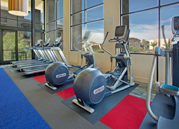 Bright gym interior here at Escape Apartments with several treadmills and ellipticals on colorful mats. Large windows show a scenic urban view, creating an inviting atmosphere.
