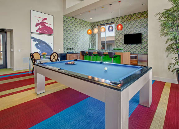 Modern game room here at Escape Apartments with a blue pool table, colorful striped carpet, abstract art, and a bar area. Pendant lights add warmth; a bamboo plant is in the corner.