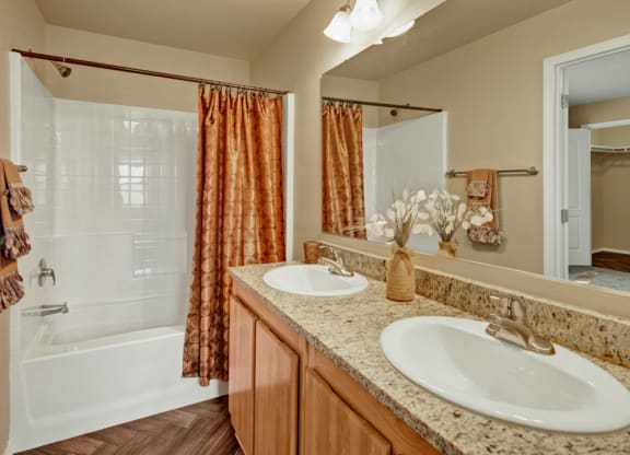 Bathroom here at Four Seasons Apartments with granite countertop, double sinks, and decorative vases. A white tub-shower with an orange curtain is in the background, conveying warmth.