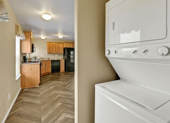 Stacked white washer and dryer in a hallway with a view of a kitchen. The kitchen here at Four Seasons Apartments features wooden cabinets, chevron-patterned flooring, and black appliances, conveying a cozy, modern feel.