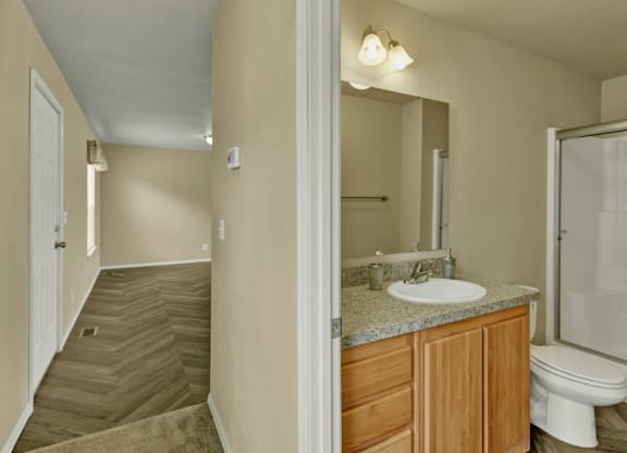 Hallway here at Four Seasons Apartments with wood-patterned floor leading to bathroom. Bathroom has a granite-topped wooden vanity, oval mirror, and shower. Walls are beige, creating a calm atmosphere.