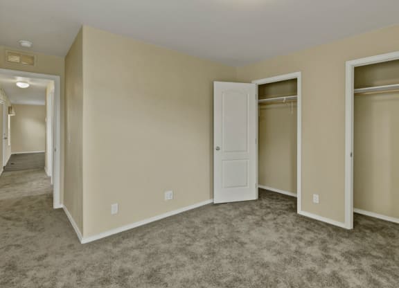 Bedroom here at Four Seasons Apartments with beige walls, open closet doors, and gray carpet. A hallway is visible leading to another lit room, creating a neutral and vacant atmosphere.