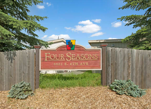 Wooden sign reading "Four Seasons, 5901 E. 6th Ave" surrounded by a wooden fence, green shrubs, and pine trees under a clear blue sky.