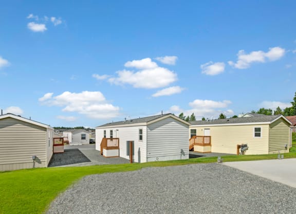 A row of mobile homes here at  Four Seasons Apartments with wooden decks is set on a neatly maintained grassy and gravel path under a bright blue sky with scattered clouds.