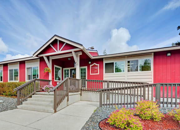 Front view of a red and white building here at The Glen Apartments, identified as a Leasing Center. The entrance features steps and a ramp, surrounded by colorful landscaping under a partly cloudy sky.