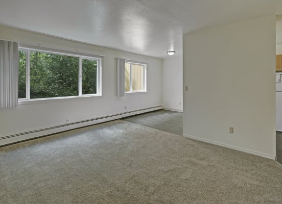 A bright living room here at The Glen Apartments with beige carpet and a large window showing green trees. White walls, partial view of a kitchen with a fridge. Calm, open atmosphere.