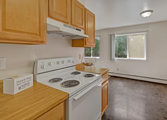 Simple kitchen here at The Glen Apartments with light wood cabinetry, white stove, and box labeled “Snack & Relax.” Bright window, empty fruit bowl, and clean, minimalist decor.
