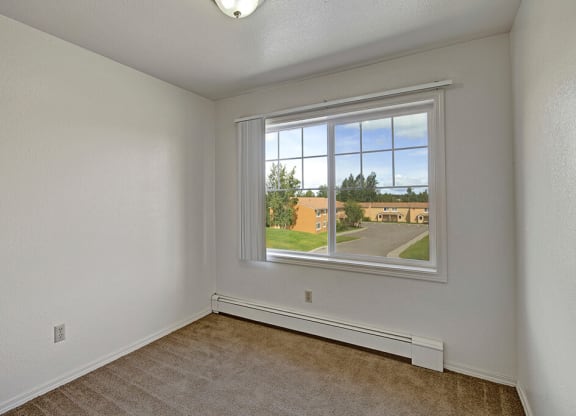 Bedroom here at The Glen Apartments with white walls, carpeted floor, and a large window revealing a view of houses, greenery, and a blue sky. Calm and clean atmosphere.