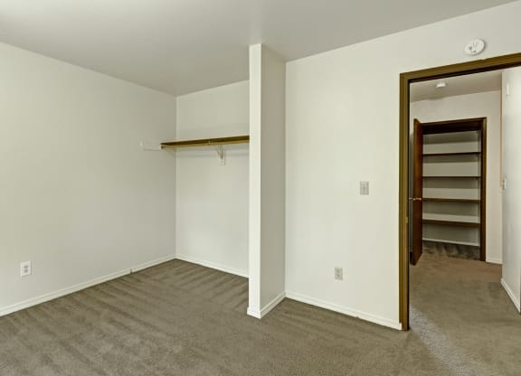 Empty closet here at The Glen Apartments with beige carpet and white walls, featuring a single wooden rod. An open door on the right leads to a room with shelving. Minimalist and clean.