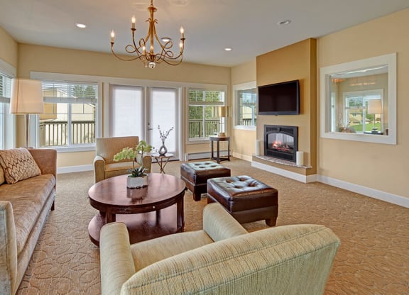 Cozy living room here at The Glen Apartments with a modern design. Features beige couches, a round wooden table, a fireplace, and large windows. A chandelier adds elegance.