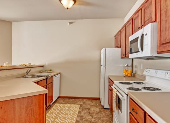 Warm kitchen here at Grand River Estates Apartment Homes with wood cabinets, white appliances, and beige countertops. A jar of oranges and neatly arranged dishware add a cozy, inviting feel.