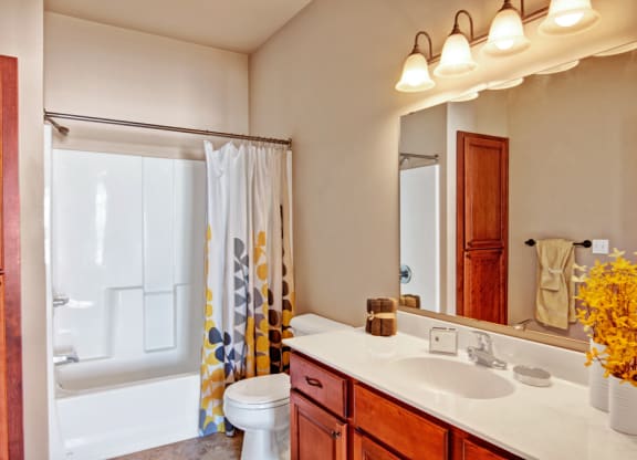 Bright bathroom here at Grand River Estates Apartment with wooden cabinets, a white counter, and double sink. Yellow accents include a vibrant shower curtain and flowers, under soft lighting.