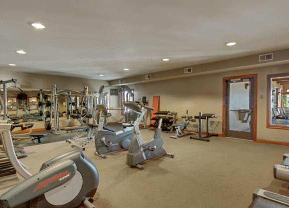 A spacious gym here at Grand River Estates Apartment Homes with various exercise equipment including treadmills, stationary bikes, weights, and mirrors. The room is well-lit and organized, creating an inviting atmosphere.