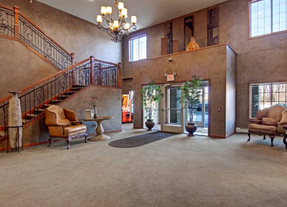 Spacious, elegant lobby here at Grand River Estates Apartment Homes with soft lighting, featuring a grand staircase, leather chairs, round tables, and large windows. Neutral tones create a warm, inviting ambiance.