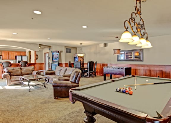 Luxurious game room here at Grand River Estates Apartment Homes featuring a pool table with cues and balls, leather sofas, foosball table, dartboard, arcade game, and warm ambient lighting.