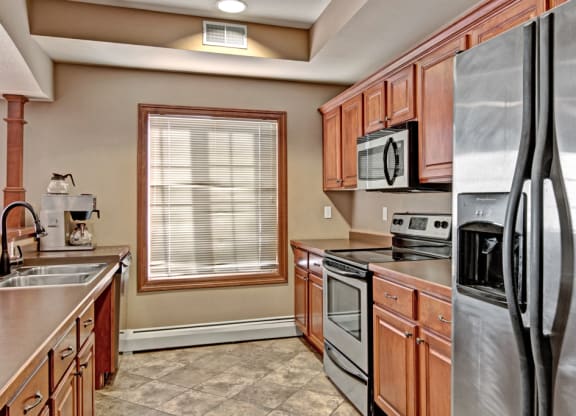 A modern kitchen here at Grand River Estates Apartment Homes with wooden cabinets, stainless steel appliances, and tiled floor. Sunlight streams through a window, creating a warm, inviting ambiance.