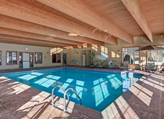 Indoor pool here at Grand River Estates Apartment Homes with clear water and blue tiles, set in a cozy room with wooden beams. Sunlight streams through large windows, casting reflections on the water.
