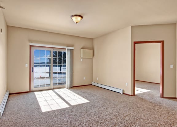 Bright, empty room here at Grand River Estates Apartment Homes with beige walls and carpet, large sliding glass door letting in sunlight, and open doorway connecting to another room. Serene and spacious.