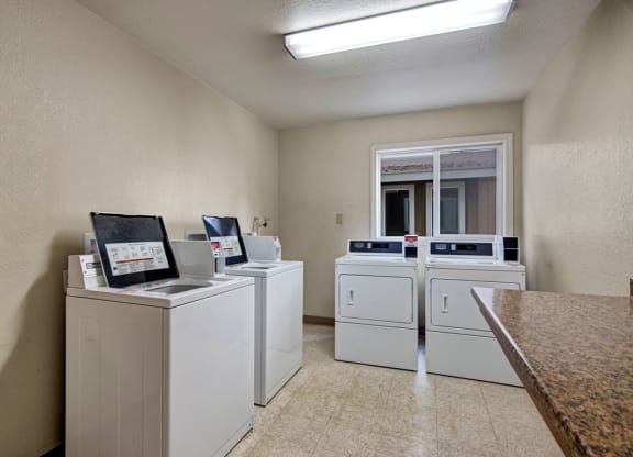 A clean, brightly-lit laundry room here at Greenbriar Apartments with four white washing machines and dryers lined against cream-colored walls. A window reflects natural light.