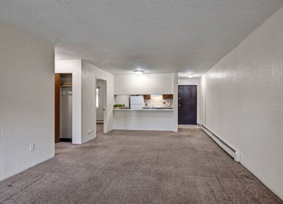 Empty living room here at Greenbriar Apartments with beige carpet and white walls, featuring a small closet on the left, a kitchen with a counter in the middle, and a dark wooden door on the right. Bright overhead lighting creates a clean, spacious feel.