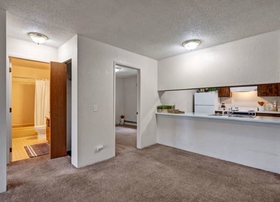Cozy apartment interior here at Greenbriar Apartments with beige carpet and white walls. Features a small kitchen with a breakfast bar, bathroom with shower curtain, and softly lit rooms.
