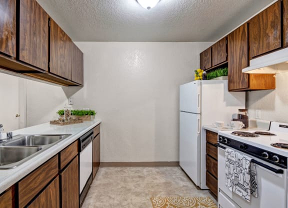 Compact kitchen here at Greenbriar Apartments with dark wood cabinets, white appliances including a fridge and stove, dual sink, and decorative green plants. Cozy and functional.