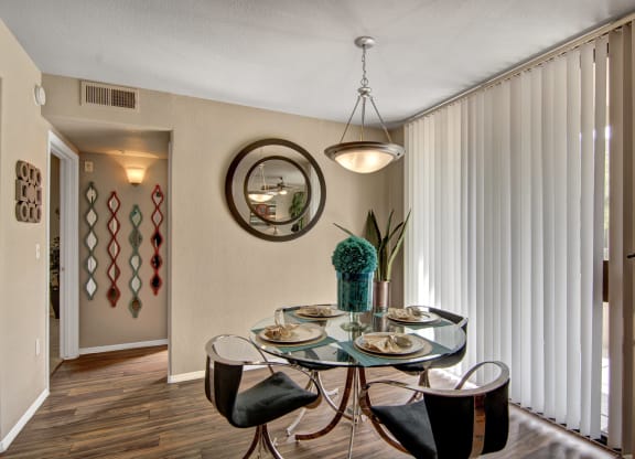 Modern dining area here at The Hawthorne Apartments with a round glass table set for four, elegant chairs, and a teal centerpiece. Wall mirror and vertical blinds create a warm, cozy ambiance.