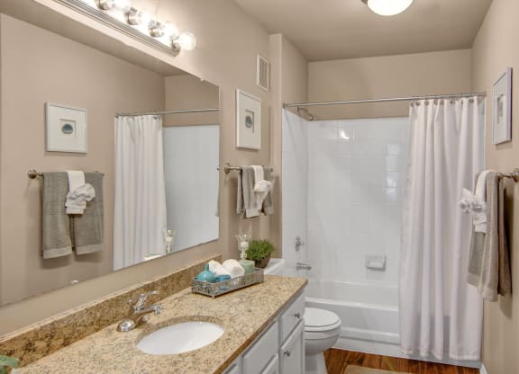 A modern bathroom here at The Hawthorne Apartments with beige walls, featuring a granite countertop, large mirror, white sink, and shower with white tiles and curtain. Fresh towels are neatly arranged.