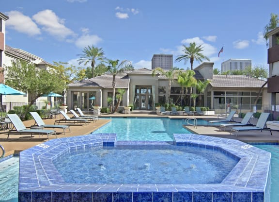 Luxurious pool area here at The Hawthorne Apartments with blue jacuzzi in the foreground, surrounded by lounge chairs. Palm trees and a modern building create a serene, sunny atmosphere.