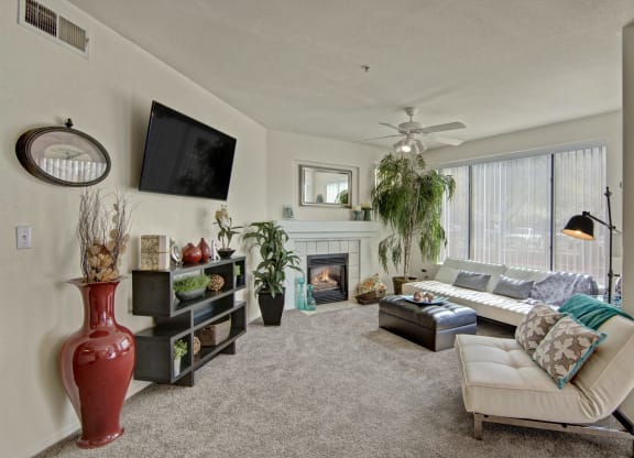Cozy living room here at The Hawthorne Apartments with neutral tones, featuring a cushioned lounge chair, large L-shaped sofa, fireplace, plants, wall-mounted TV, and decorative items.