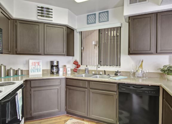 Modern kitchen here at The Hawthorne Apartments with dark wood cabinets, beige countertops, and stainless steel appliances. Decor includes a "Home Sweet Apartment" sign and red mugs.