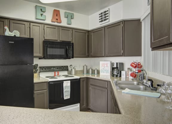 Modern kitchen here at The Hawthorne Apartments with dark cabinets, black appliances, and beige countertops. Decor includes colorful "EAT" letters and a rooster figure, creating a cozy vibe.