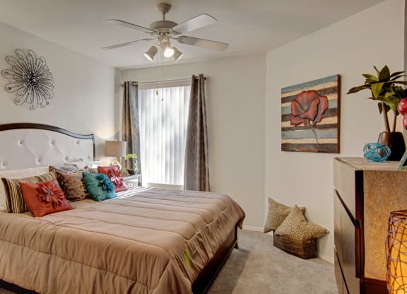 Bedroom here at The Hawthorne Apartments with a neutral tone features a large bed with colorful pillows. A flower painting and metal wall art add warmth, while soft light from a lamp enhances coziness.
