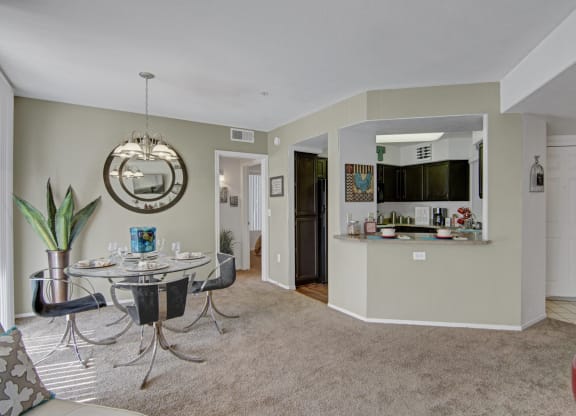 Modern dining area here at The Hawthorne Apartments with a glass table, set for four, black chairs, and a large mirror on a pale green wall. Adjacent kitchen with dark cabinets. Cozy and inviting atmosphere.