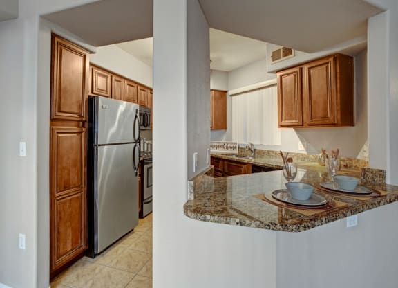 Modern kitchen here at The Hawthorne Apartments with stainless steel fridge, brown cabinets, and granite countertops. A breakfast bar with two place settings adds a cozy touch.