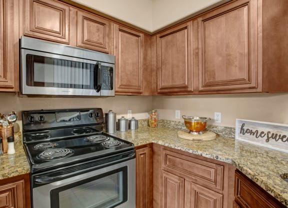 Modern kitchen here at The Hawthorne Apartments with brown wooden cabinets, granite countertops, and stainless steel appliances. Includes a stove, microwave, jars, utensils, and "home sweet" sign.